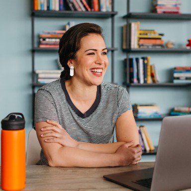 A smiling woman sits at a desk with a laptop and orange water bottle.