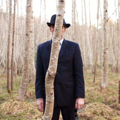 A man standing behind a small tree trunk.