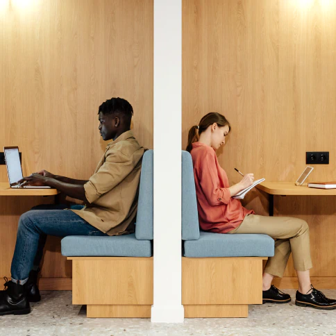 Two people sitting in separate back-to-back booths. They are working on their devices.