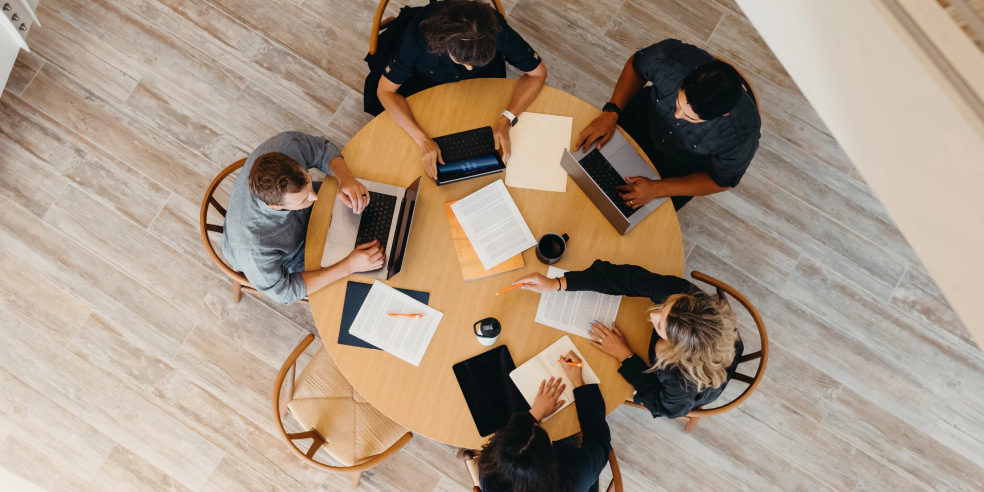 A group of people working together at a round table with laptops and papers.