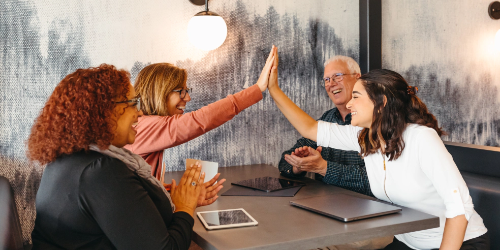 A small team is seated around a table. Everyone is smiling. Two people are high-fiving each other.