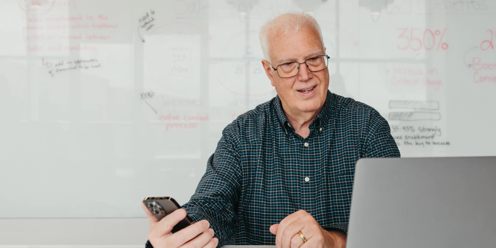 A man holds a cell phone while looking a computer screen.